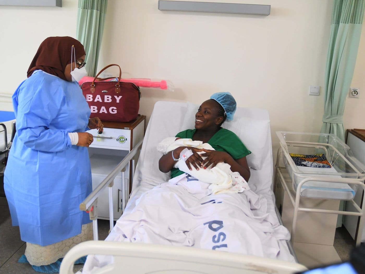 The President, in a surgical gown and mask, greeting a mother resting with her newborn in a maternity ward