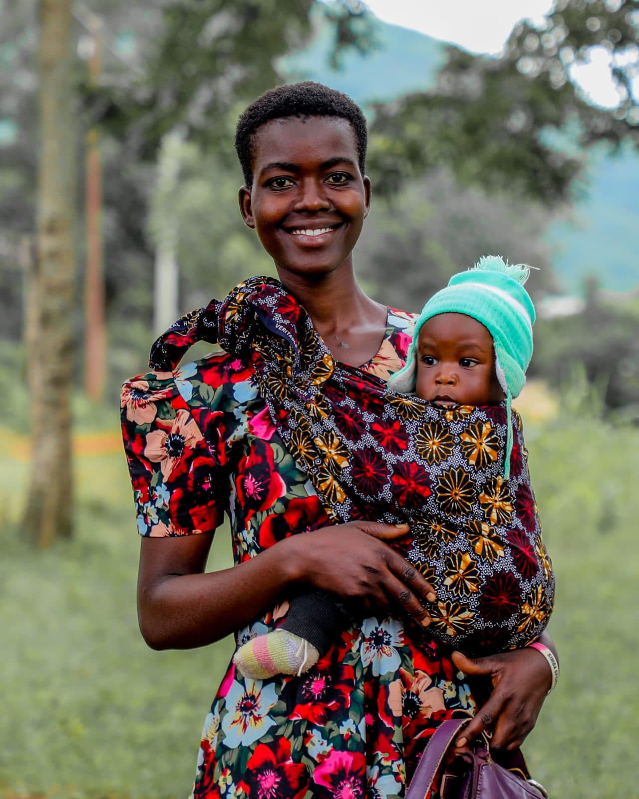 A mother smiling at the camera with her baby carried in a patterned cloth sling