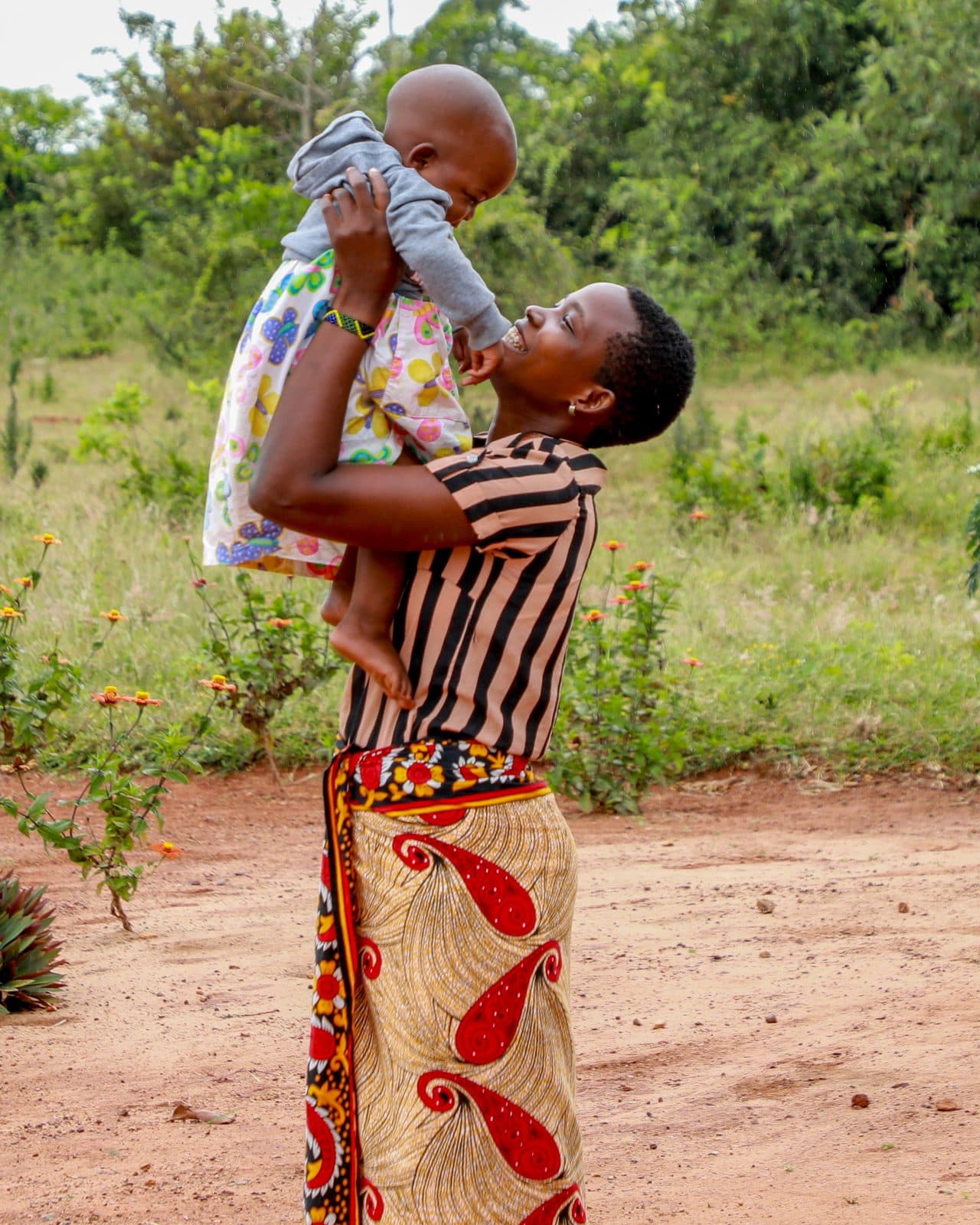 A mother lifting her baby above her head, both laughing, in a green open space