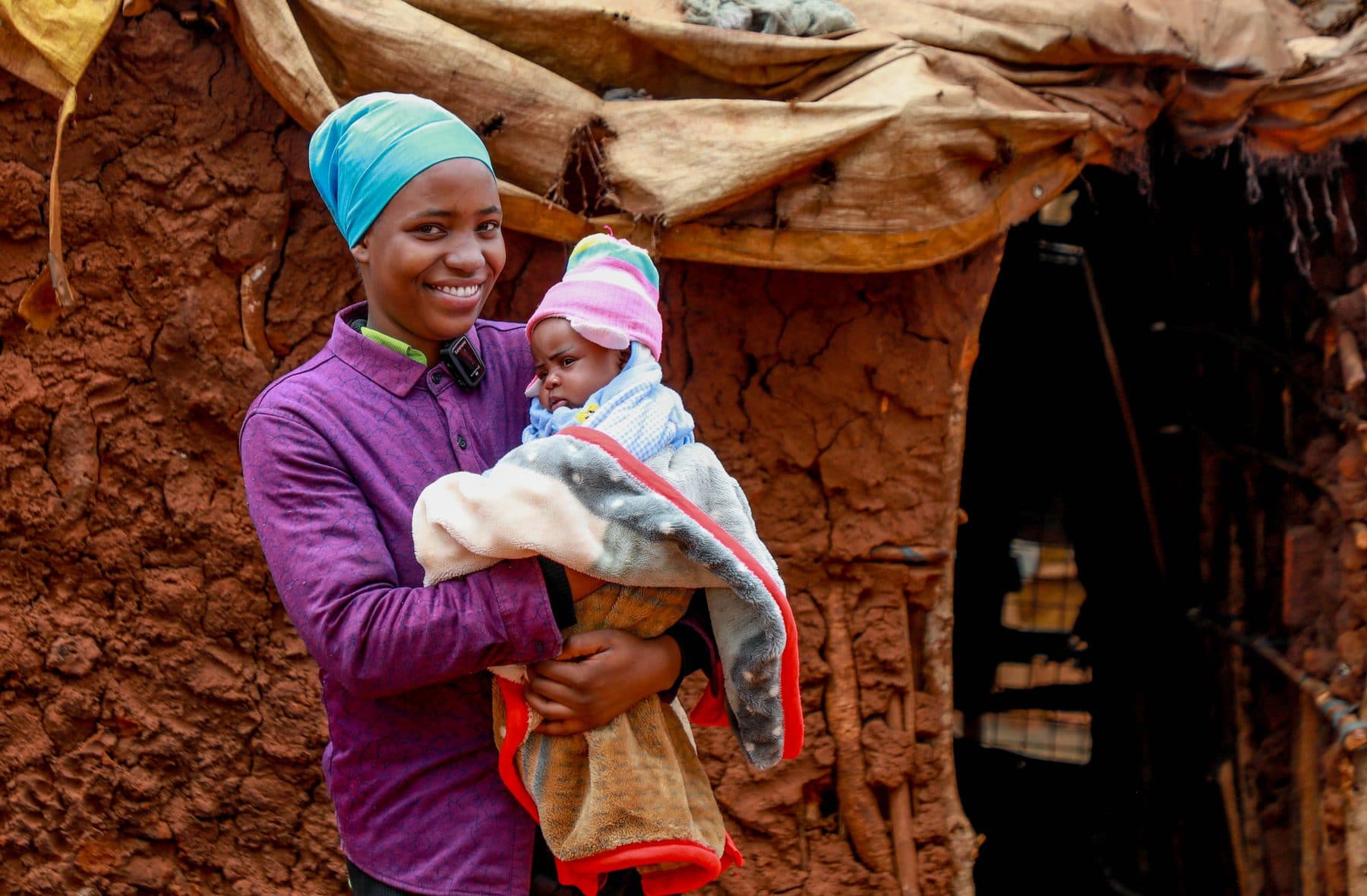 A mother standing outside her home holding her wrapped newborn, smiling at the camera
