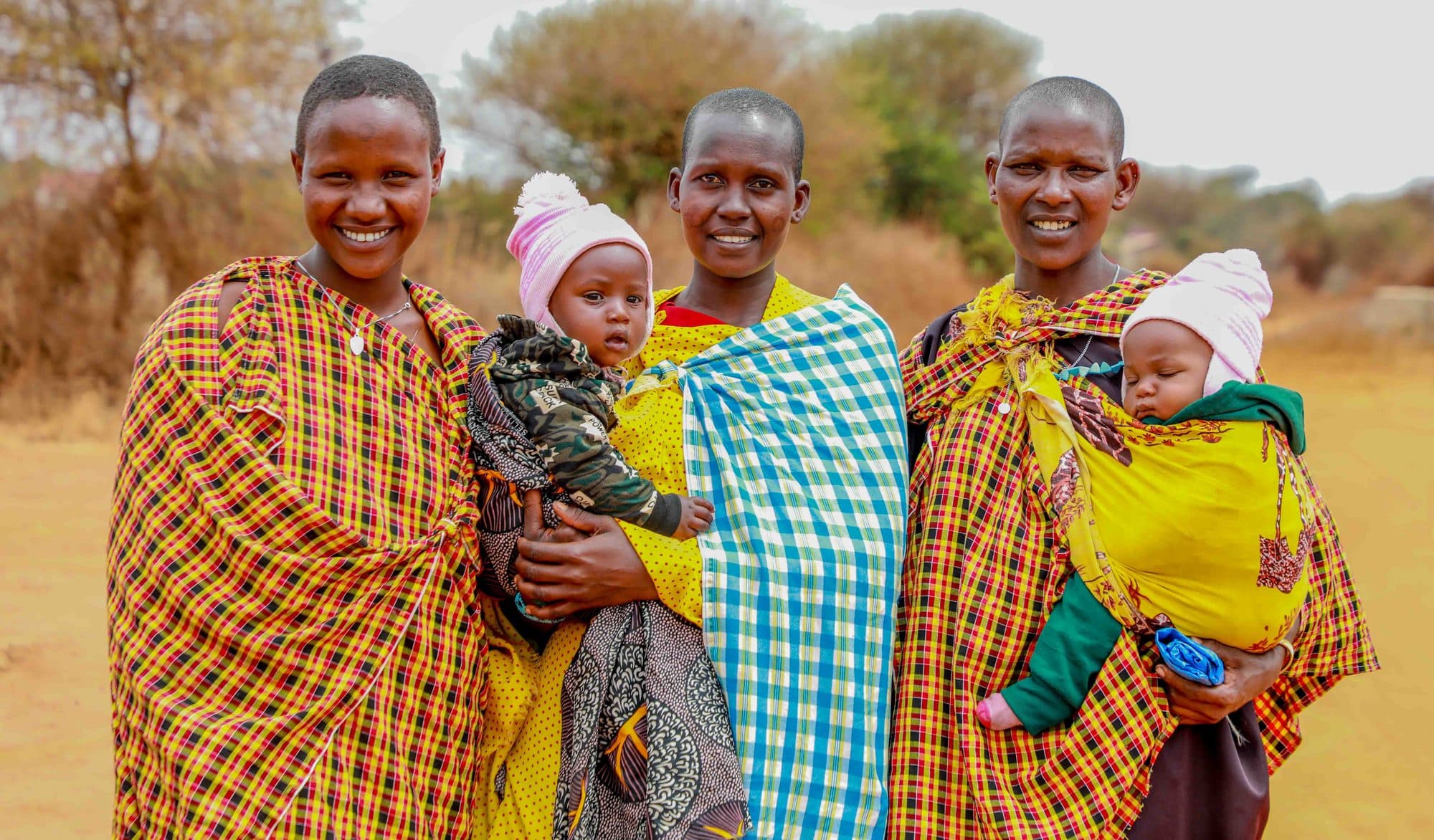 Three mothers standing together outdoors, each carrying a baby, all smiling at the camera